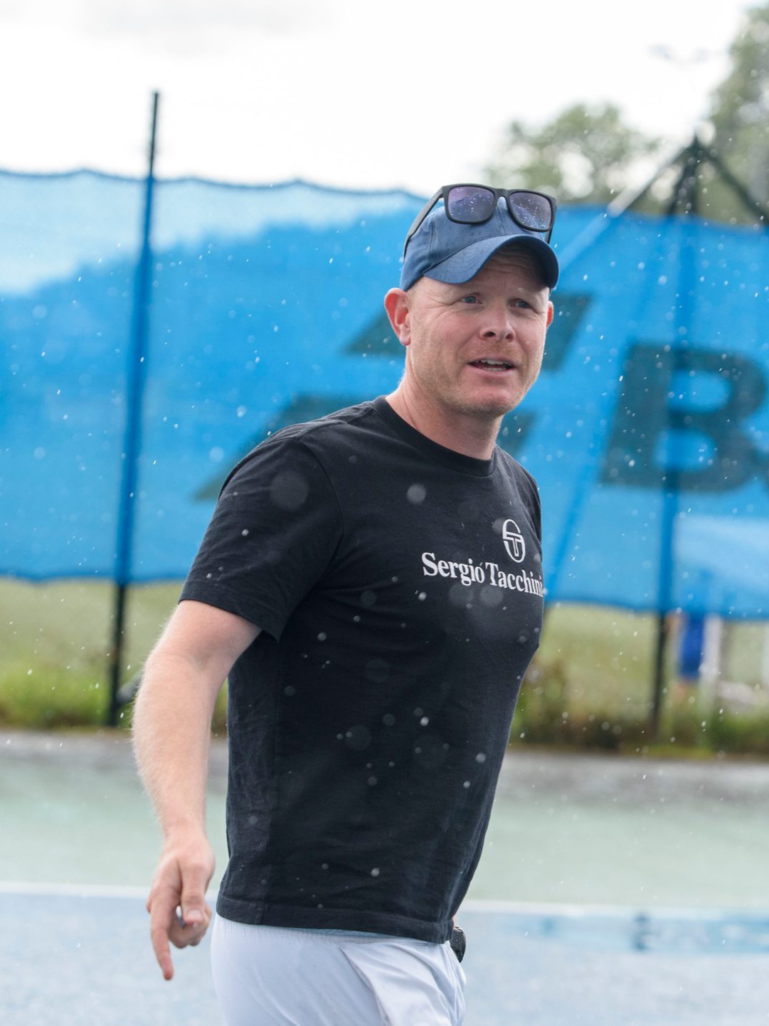 Henrik i svart t-skjorte og blå cap på tennisbane i regnvær